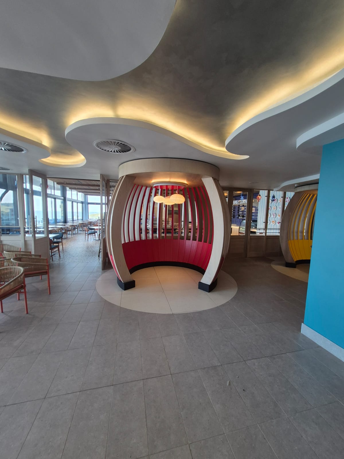A red slatted cocoon booth on a raised terrazzo plinth under a curved cove-lit ceiling